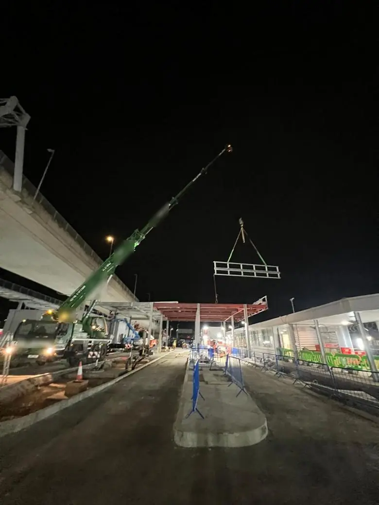Mobile crane lifting a steel frame at night beside a flyover